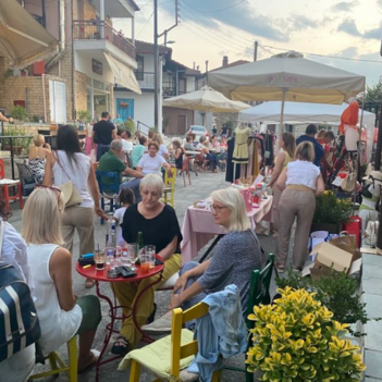 Υπαίθριο bazaar με πολύχρωμους πάγκους γεμάτους χειροποίητα προϊόντα, ρούχα, κοσμήματα και τοπικές λιχουδιές. Επισκέπτες περπατούν και εξερευνούν την αγορά σε ζωντανή, γιορτινή ατμόσφαιρα